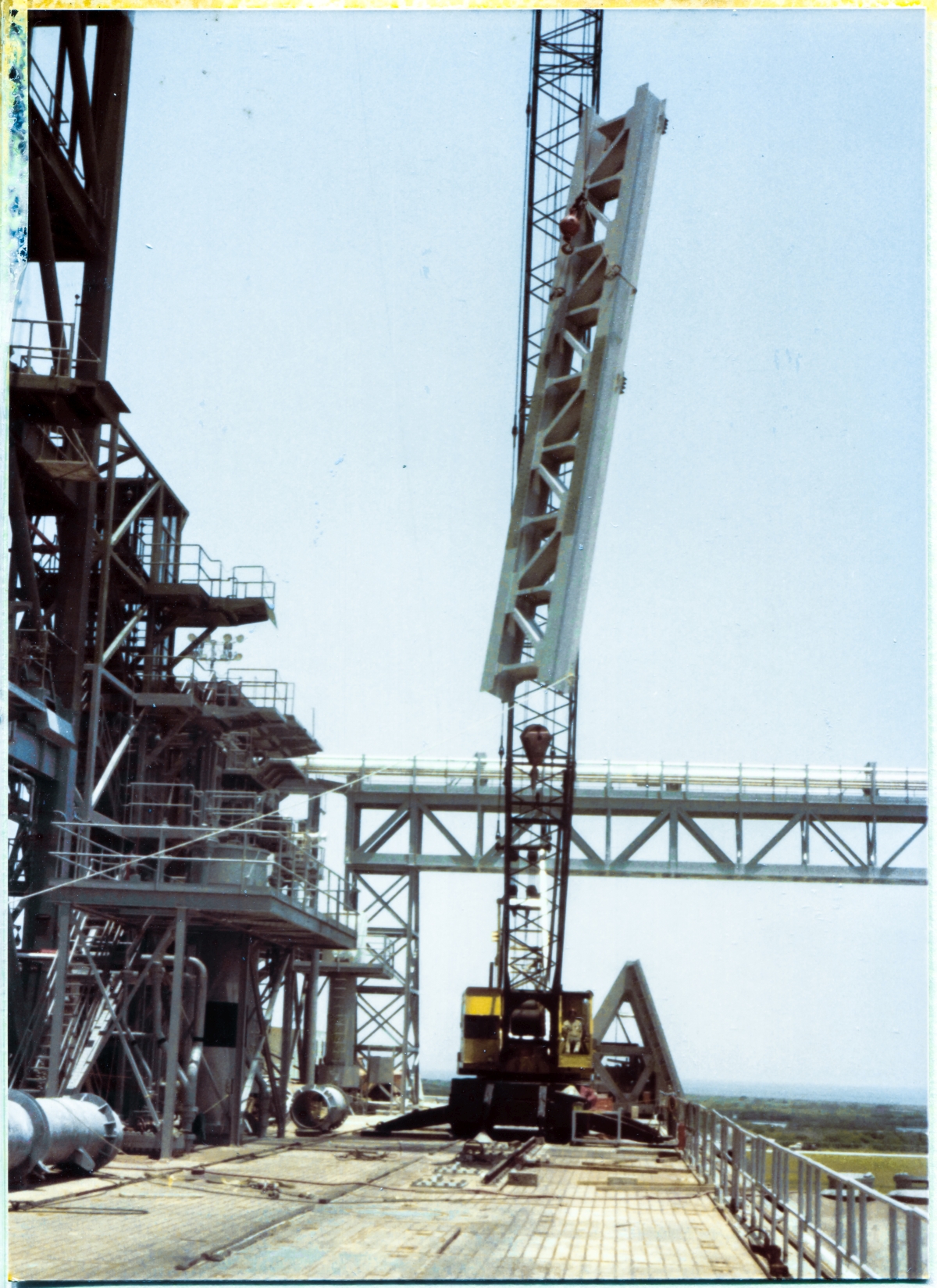 Image 074. The GOX Arm Strongback now begins its vertical journey to the top of the Fixed Service Structure (a small part of which is visible along the left margin of the photograph, extending up to and beyond the top) at Space Shuttle Launch Complex 39-B, Kennedy Space Center, Florida. Beneath it, on the ground, all personnel have been cleared from the area, and will remain so as a safety precaution until the Strongback is firmly welded to the FSS Primary Framing, far above where it is located at this moment. A Union Ironworker from Local 808, working for Ivey Steel, remains unseen, handling the tag line which is visible, attached to the lower right-side corner of the Strongback, keeping it properly oriented as it rises higher and higher into the sky. Photo by James MacLaren.